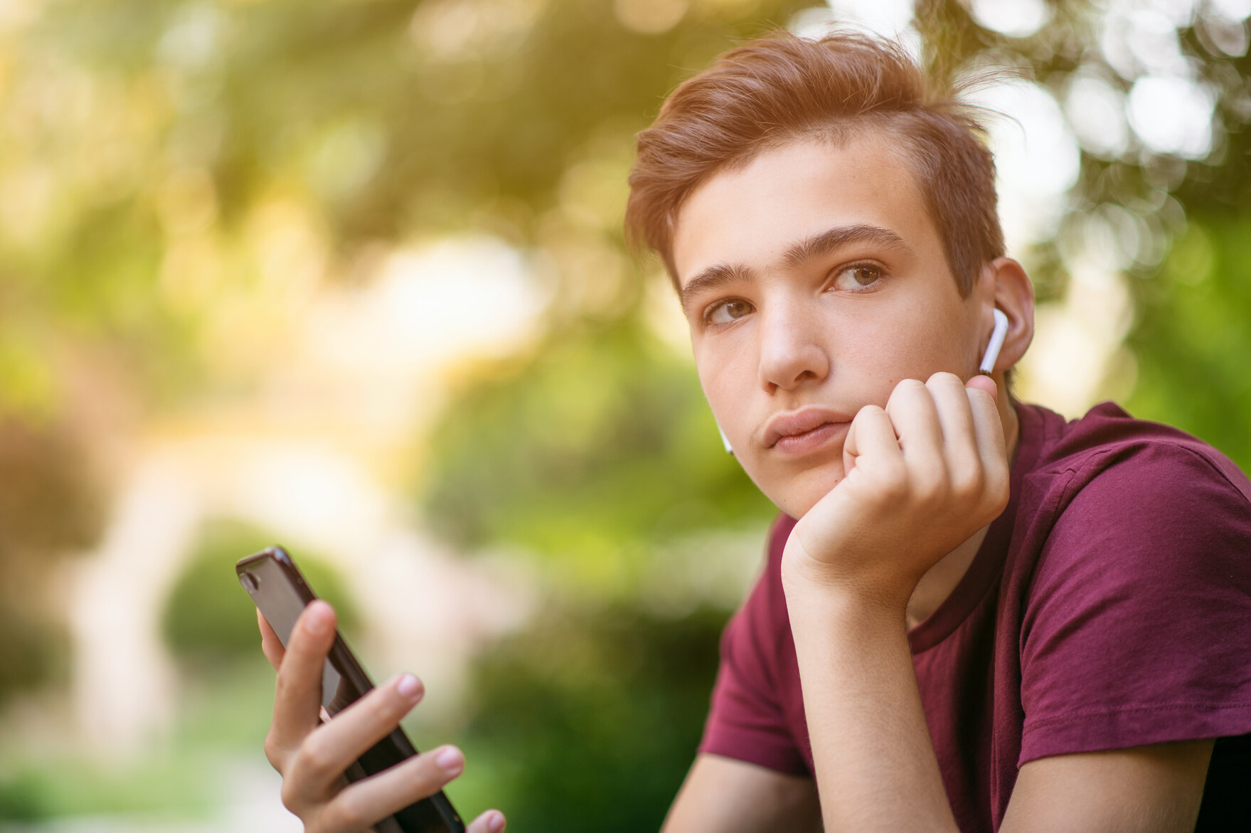 ein Junge sitzt im Park, hält sein Handy in der Hand und schaut nachdenklich 