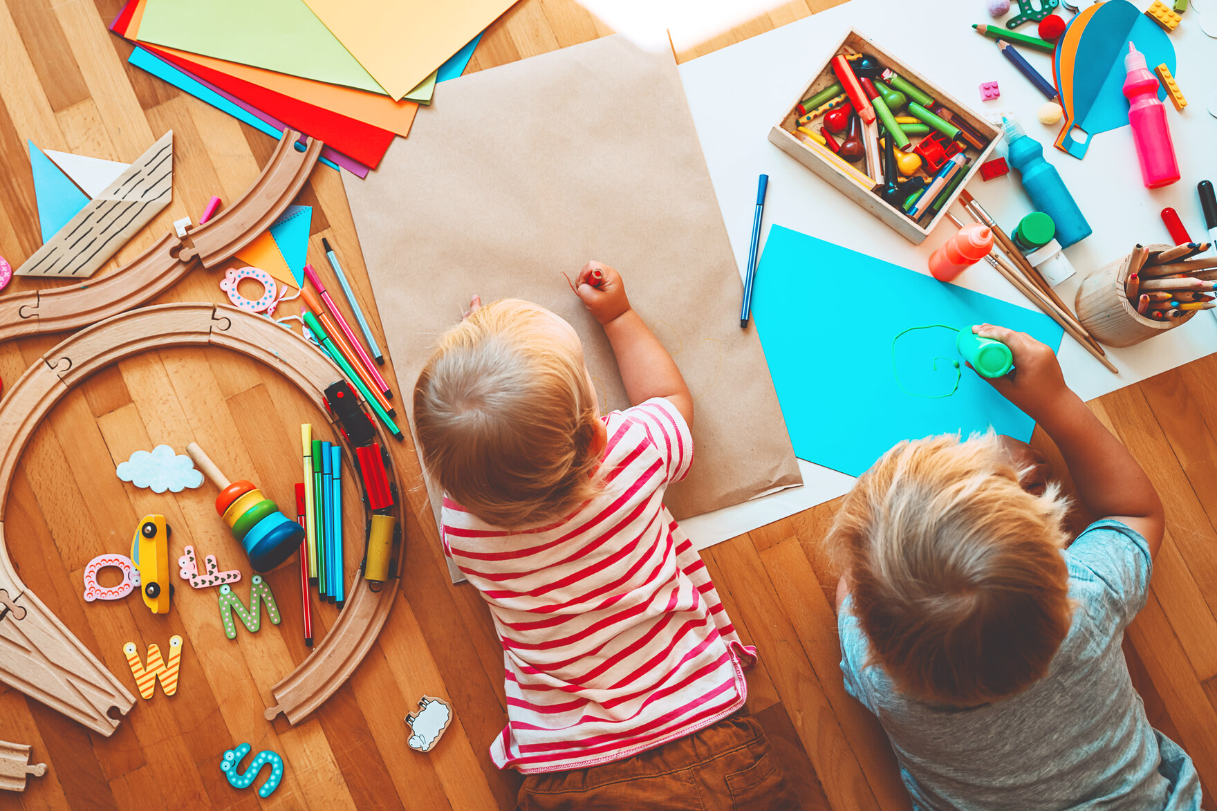 Zwei Kinder liegen auf dem Laminat zwischen Spielzeug und malen
