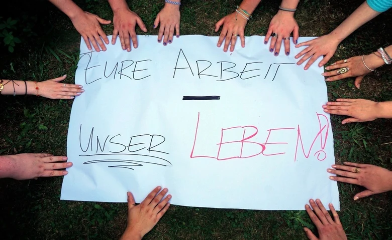 Das Bild zeigt ein weißes Plakat, auf dem „EURE ARBEIT - UNSER LEBEN!“ steht. Viele Hände umringen das Plakat, symbolisch für Gemeinschaft und den Ausdruck einer Botschaft oder Forderung.