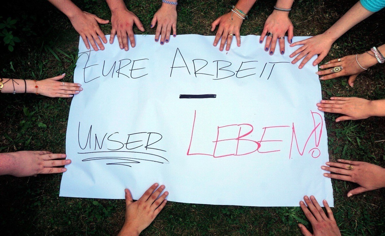 Das Bild zeigt ein weißes Plakat, auf dem „EURE ARBEIT - UNSER LEBEN!“ steht. Viele Hände umringen das Plakat, symbolisch für Gemeinschaft und den Ausdruck einer Botschaft oder Forderung.