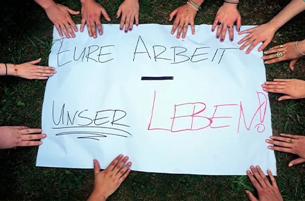 Das Bild zeigt ein weißes Plakat, auf dem „EURE ARBEIT - UNSER LEBEN!“ steht. Viele Hände umringen das Plakat, symbolisch für Gemeinschaft und den Ausdruck einer Botschaft oder Forderung.