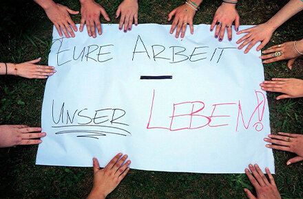 Das Bild zeigt ein weißes Plakat, auf dem „EURE ARBEIT - UNSER LEBEN!“ steht. Viele Hände umringen das Plakat, symbolisch für Gemeinschaft und den Ausdruck einer Botschaft oder Forderung.